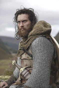 a man with long hair and beard wearing armor sitting on the ground in front of mountains