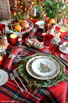 the table is set for christmas dinner with red and white dishes, silver utensils, oranges, pine cones, and greenery