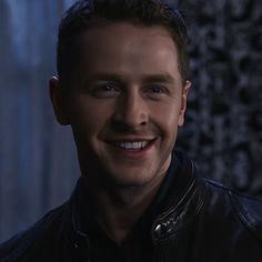 a smiling man wearing a leather jacket in the dark with his eyes wide open and looking at the camera