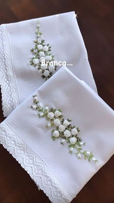 two white napkins with flowers on them sitting on top of a wooden table next to each other