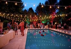 people standing around a pool at night with lights strung over the pool and in the background