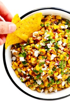 a hand holding a tortilla chip over a bowl of mexican corn salad