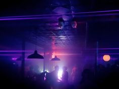 people are dancing in a dark room with lights on the ceiling and purple lighting hanging from the ceiling