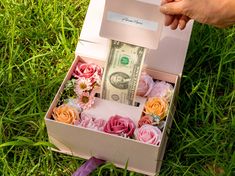 a person is holding a money bill in a box with flowers on the grass behind it