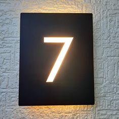 a black square sign with the number seven lit up on it's side against a white brick wall