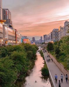 people are walking on the sidewalk next to a river and buildings in the background at sunset