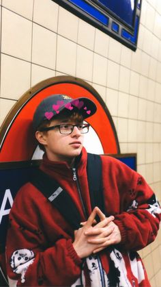 a young man wearing glasses and a red jacket standing in front of a subway sign