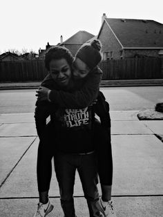 two people are hugging each other on the sidewalk in front of a house and one person is wearing a beanie