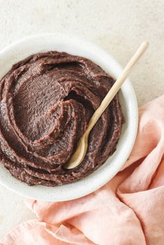 a white bowl filled with chocolate frosting on top of a pink napkin next to a wooden spoon