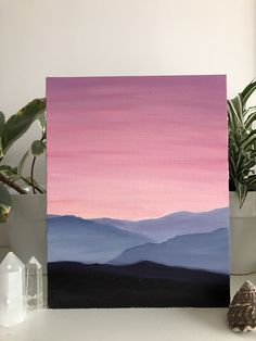 an acrylic painting of mountains in pink and blue on a white table next to potted plants