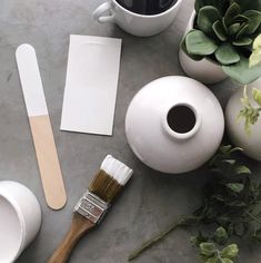 a table topped with white vases filled with plants and paint brushes next to a cup of coffee