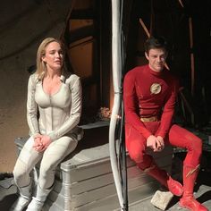 two people dressed in costumes sit on a bench outside an abandoned building, one man is wearing a red suit and the other wears a white shirt