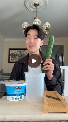 a man holding a cucumber in front of a cup and container on top of a table