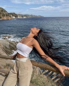 a woman standing on top of a wooden rail next to the ocean with her hair blowing in the wind