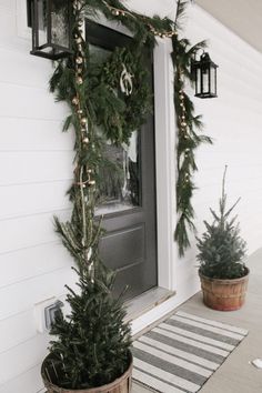 two potted christmas trees sitting on the front porch next to a door with lights