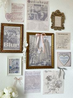 a wall covered in pictures and photos with flowers on top of it, next to a vase filled with white roses