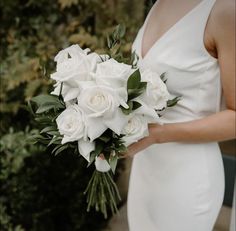 a woman in a white dress holding a bridal bouquet with roses and greenery