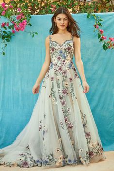 a woman standing in front of a blue backdrop wearing a dress with flowers on it