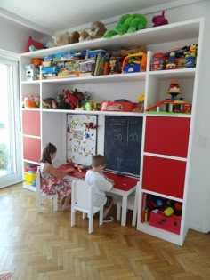 two children are sitting at a desk in front of a bookcase with toys on it