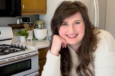 a woman sitting in front of an oven smiling at the camera with her hand on her chin