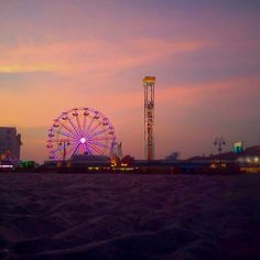 a ferris wheel sitting on top of a sandy beach next to tall buildings at dusk