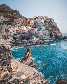 a woman sitting on top of a cliff next to the ocean with buildings in the background