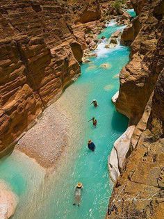 several people are swimming in the blue water near some rocks and cliffs, while another person is standing on one side of the river