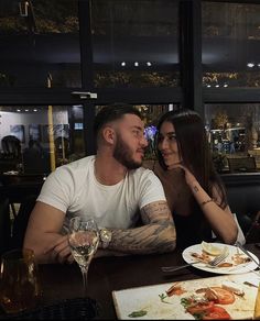 two people sitting at a table with food and drinks in front of them, one man has his hand on the woman's face