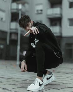 a young man crouching down on the ground pointing at something with his hand while wearing white sneakers