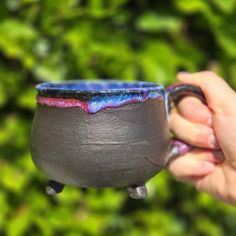 a hand holding a black and blue cup with purple glaze on the outside, in front of green foliage