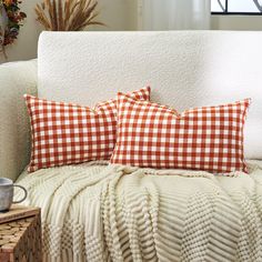 two orange and white plaid pillows on a couch next to a basket with coffee mugs