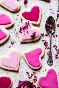 heart shaped sugar cookies with pink icing and rose petals on a baking sheet next to a spoon