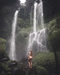 a woman standing in front of a waterfall with her back turned to the camera,