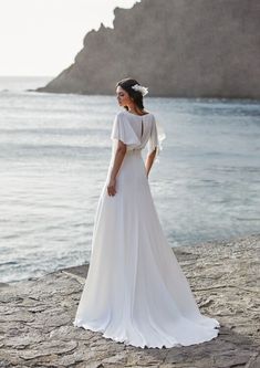 a woman in a white wedding dress standing on rocks near the water with her back to the camera