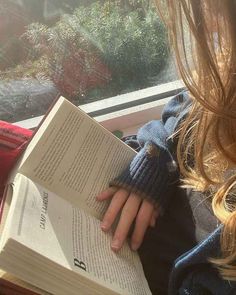 a woman reading a book while sitting on a bench in front of a window and looking out the window