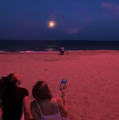 two women sitting on the beach looking at their cell phones in front of the moon