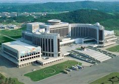 an aerial view of a large building in the middle of a green area with mountains in the background