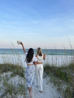 two women are walking on the beach with their arms in the air and one is holding something up