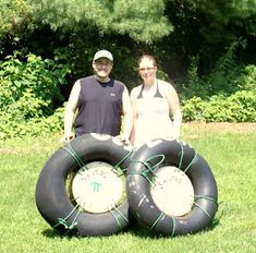 two people standing next to each other in the grass with large black tires on them