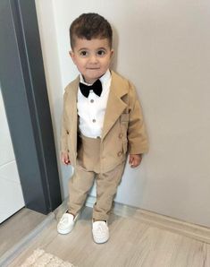 a little boy in a suit and bow tie standing next to a wall wearing white shoes