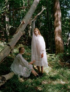 two women sitting on the ground next to each other in the woods, one wearing a white dress