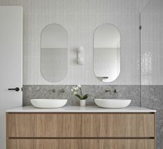 two white sinks sitting on top of a wooden cabinet next to mirrors and a vase with flowers