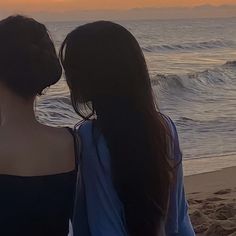 two women are standing on the beach looking at the ocean and sunset in the background