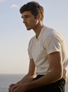 a man sitting on top of a wooden bench next to the ocean with his hands in his pockets