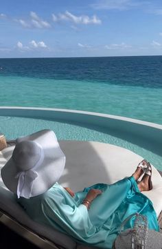 a woman laying on top of a white couch next to the ocean and wearing a hat