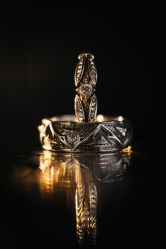 an intricately designed wedding ring sits on a reflective surface with its reflection in the water