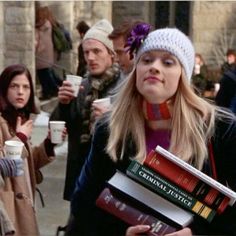 a woman holding books and drinking coffee while walking down the street with other people behind her