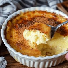 a hand holding a spoon over a baked dessert in a white dish on a wooden table