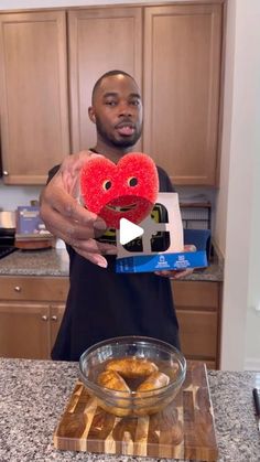 a man holding up a heart shaped doughnut in front of a bowl of donuts