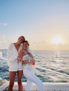 two beautiful women standing on the back of a boat in front of the ocean at sunset